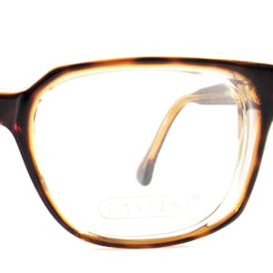 May include: Close-up of eyeglasses with a brown tortoiseshell frame. The glasses have a rectangular shape with rounded edges and clear lenses. The temples are a light brown color. The brand name "LASTIS" is visible on the lens.