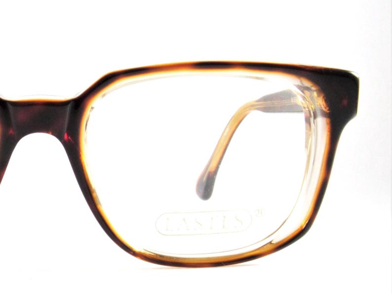 May include: Close-up of eyeglasses with a brown tortoiseshell frame. The glasses have a rectangular shape with rounded edges and clear lenses. The temples are a light brown color. The brand name "LASTIS" is visible on the lens.