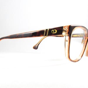 May include: Brown and tan eyeglasses with a tortoiseshell pattern on the temples. The frames have a rectangular shape with clear lenses. A gold accent and the letter "D" are visible on the temple.