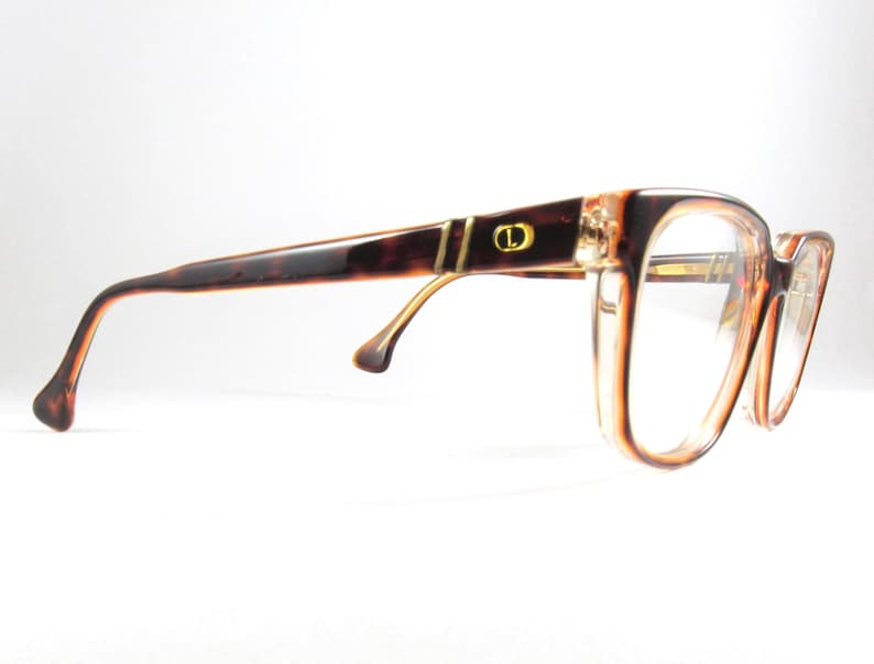 May include: Brown and tan eyeglasses with a tortoiseshell pattern on the temples. The frames have a rectangular shape with clear lenses. A gold accent and the letter "D" are visible on the temple.