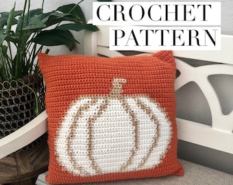 May include: A crocheted pillow with a white pumpkin design on an orange background. The pillow is sitting on a white bench.