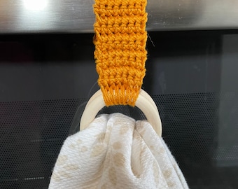May include: Mustard yellow crochet towel holder with a wooden ring, holding a white and beige patterned towel. The holder is attached to a stainless steel appliance. The towel has a leaf pattern.