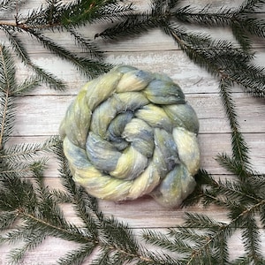 Puede incluir: Un ovillo de hilo hilado a mano en tonos amarillo pálido, azul y blanco. El hilo está dispuesto en forma circular, con un aspecto texturizado. El fondo es una superficie de madera clara, con ramas de pino verdes en las esquinas.