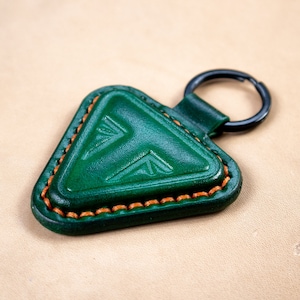 May include: A green leather triangle keychain with a decorative pattern and orange stitching. The keychain has a black metal ring.