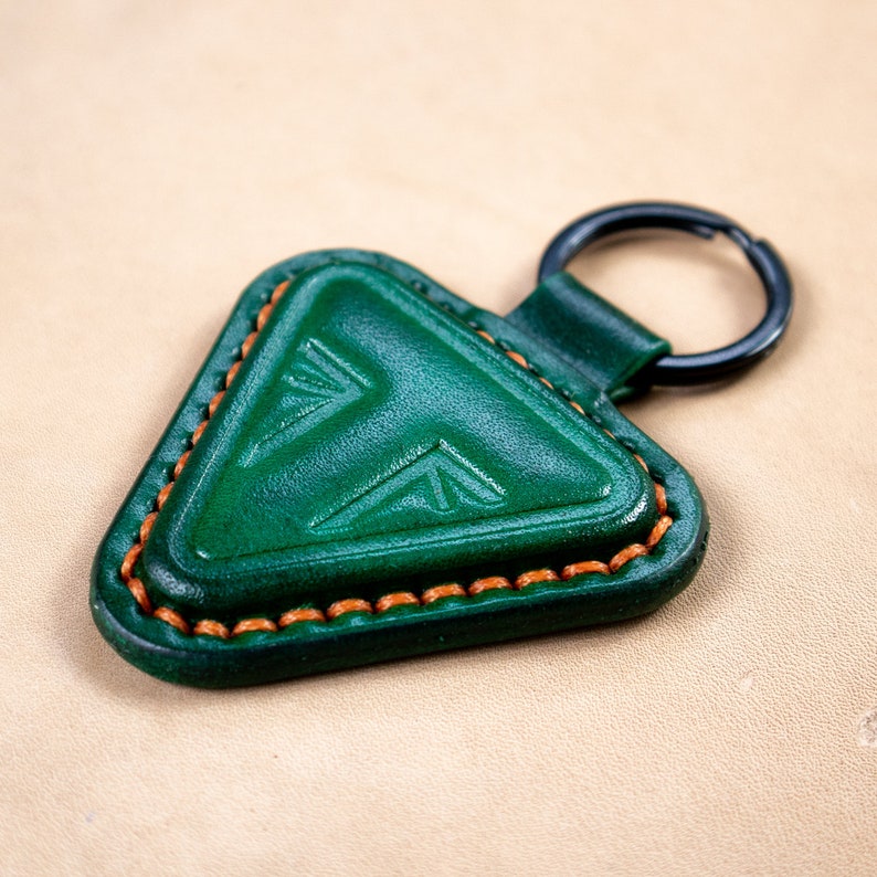 May include: A green leather triangle keychain with a decorative pattern and orange stitching. The keychain has a black metal ring.