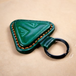 May include: A green leather keychain with a triangular shape and a black metal ring. The keychain has a decorative stitch around the edge.