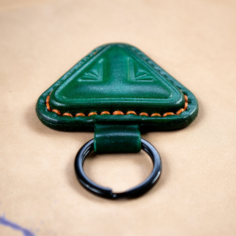 May include: A green leather triangle-shaped keychain with a black metal ring. The keychain has a decorative stitch around the edge and a small design in the center.