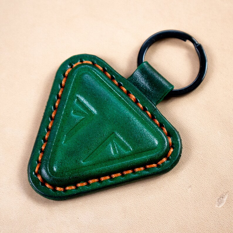 May include: A green leather keychain with a triangle-shaped charm. The charm has a stitched border and a decorative pattern. The keychain has a black metal ring.