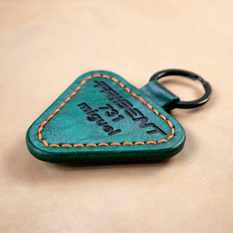 May include: A green leather keychain with a triangle shape and orange stitching. The keychain has the text "Present 731 Miguel" printed on it.