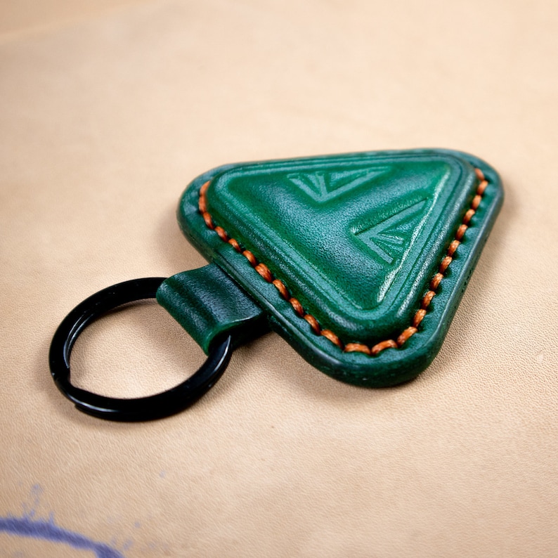 May include: A green leather key chain with a triangle-shaped tag. The tag has a decorative design and is stitched with orange thread.