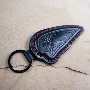 Könnte beinhalten: Schwarzer Leder-Schlüsselanhänger mit einem flügelförmigen Design und roten Nähten. Der Schlüsselanhänger hat ein geprägtes Emblem. Der Schlüsselring ist schwarz. Das Leder hat eine strukturierte Optik.