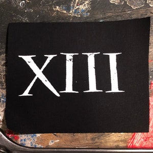 May include: A black square patch with the Roman numeral XIII in white. An adjustable wrench, labeled "Chrome Vanadium Steel 8", is positioned below the patch. The background is a weathered, multi-colored surface.