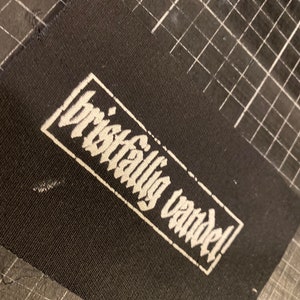 K&ouml;nnte beinhalten: Schwarzer Stoff-Patch mit wei&szlig;em Text, der in einer stilisierten Schrift "brilliantly vandal!" lautet.