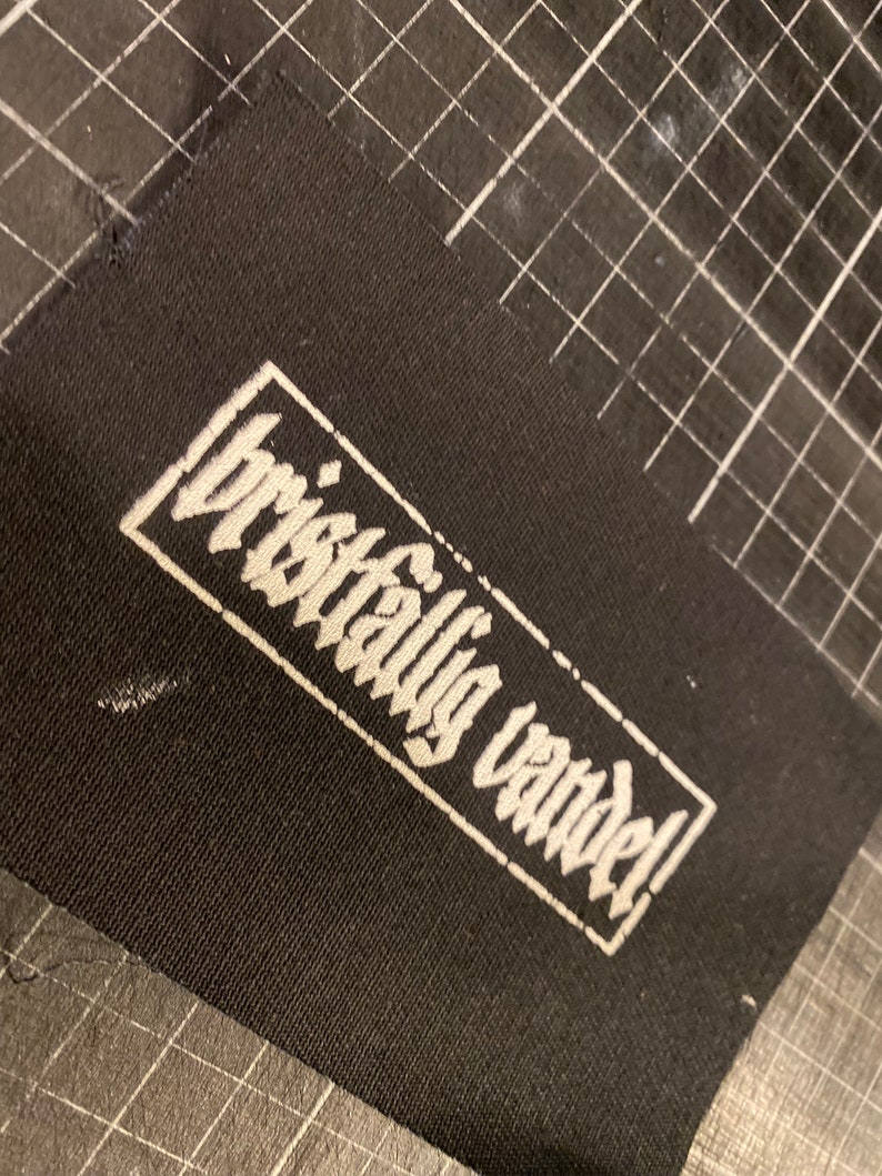 K&ouml;nnte beinhalten: Schwarzer Stoff-Patch mit wei&szlig;em Text, der in einer stilisierten Schrift "brilliantly vandal!" lautet.