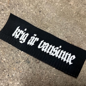Könnte beinhalten: Schwarzes Stofflabel mit weißem Text, der "krig är vansinne" lautet.