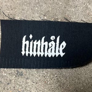 Op de afbeelding: Zwarte stoffen patch met witte tekst die "hinhaile" in een gotisch lettertype leest.