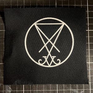 May include: Black fabric patch featuring a white sigil design within a circle. The sigil includes intersecting lines and a stylized inverted triangle. The patch is square and appears to be made of woven material.