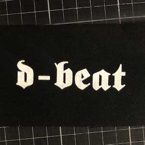 Op de afbeelding: Witte geborduurde patch met de tekst "D-beat" in gotische lettertype op een zwarte stoffen achtergrond.