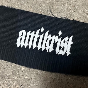 May include: Black fabric patch with the word "antichrist" printed in white in a gothic font.