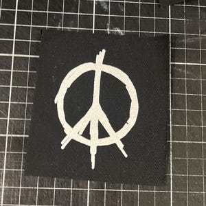 以下が含まれることがあります： 黒い布のパッチに白い平和のシンボルが描かれている
