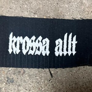 May include: Black fabric patch with white embroidered text that reads "krossa allt".