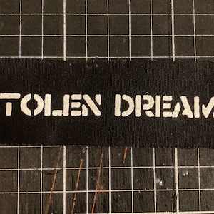 May include: A black fabric strip with the words "STOLEN DREAMS" printed in white, stencil-style lettering. The fabric is placed on a black grid surface.
