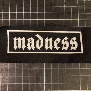 Puede incluir: Parche de tela negro con borde blanco y la palabra "madness" escrita en letras góticas blancas.