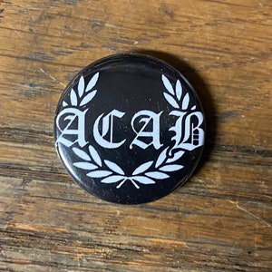 May include: A black button with a white wreath and the text "ACAB" in a white gothic font.