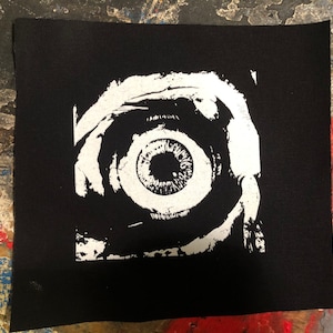 May include: A black and white patch with a large eye graphic. The eye is white with a brown iris and pupil. The eye is surrounded by a white swirl design.