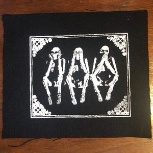 Pode incluir: Patch quadrado preto com uma ilustração branca de três esqueletos na pose 'não ver o mal, não ouvir o mal, não falar o mal'. O design é emoldurado com elementos decorativos de canto.