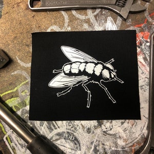 Op de afbeelding: Een zwarte strijkpatch met een witte omtrek van een vlieg. De vlieg kijkt naar rechts en heeft gedetailleerde vleugels en poten.