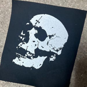 May include: Black square patch featuring a white skull design. The skull has a cross on top. The patch is made of fabric and has a raw edge. The design is a bold, high-contrast graphic.