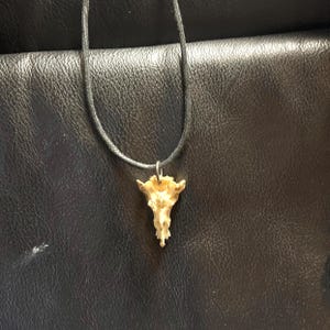 Op de afbeelding: Een zwarte koordsnoer met een kleine, lichtbruine bot hanger in de vorm van een schedel.