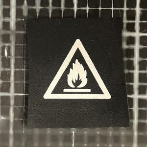 Könnte beinhalten: Ein schwarzes Stoffetikett mit einem weißen Warnsymbol. Das Symbol ist ein Dreieck mit einer Flamme, das auf eine Brandgefahr hinweist. Das Etikett ist quadratisch und hat eine strukturierte Oberfläche.