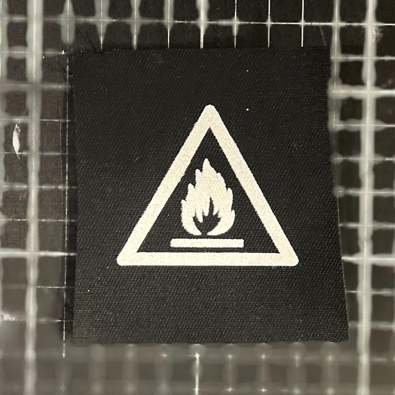 K&ouml;nnte beinhalten: Ein schwarzes Stoffetikett mit einem wei&szlig;en Warnsymbol. Das Symbol ist ein Dreieck mit einer Flamme, das auf eine Brandgefahr hinweist. Das Etikett ist quadratisch und hat eine strukturierte Oberfl&auml;che.