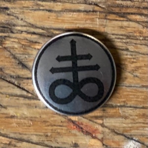 Può includere: Un bottone color argento con un pentagramma invertito nero e un simbolo di infinito.
