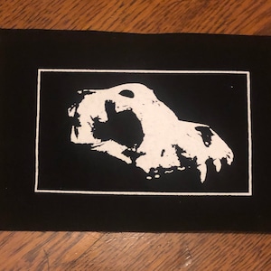 May include: Black and white patch with a white skull design. The skull is facing the right side of the image and has an open mouth with sharp teeth. The skull is surrounded by a white border.