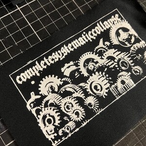以下が含まれることがあります： 黒い生地のパッチ。白のゴシック様式のフォントで「completesystematiccollapse」の文字が書かれています。パッチには、白いインターロッキングギアの詳細なイラストが描かれており、機械的なデザインになっています。パッチはおそらく衣類やアクセサリー用です。