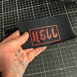 May include: Black rectangular patch with the word "HELL" in red gothic lettering. The patch is held against a black grid surface. The text is inside a red rectangular border.