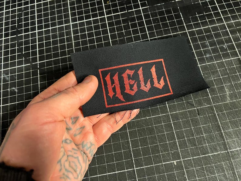 May include: Black rectangular patch with the word "HELL" in red gothic lettering. The patch is held against a black grid surface. The text is inside a red rectangular border.