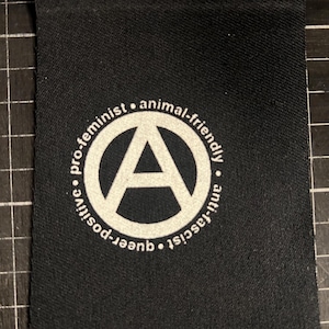 Peut inclure: Tissu noir avec un motif circulaire blanc contenant la lettre "A". Le cercle est entouré des mots "pro-féministe", "animal-friendly", "anti-fasciste", "queer" et "positive".