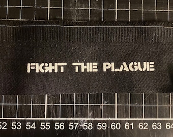 Op de afbeelding: Zwarte stoffen patch met witte tekst die "FIGHT THE PLAGUE" luidt.
