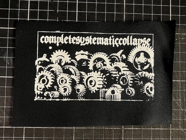 以下が含まれることがあります： 黒い生地のパッチ。白いゴシック体の「completesystematiccollapse」の文字と、連動する歯車の詳細なイラストが描かれています。 パッチは、切りっぱなしの縁です。