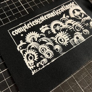 以下が含まれることがあります： 白文字で「completely systematic collapse」と書かれた黒い生地のパッチ。白い歯車が組み合わさった詳細なデザインが特徴です。衣類やアクセサリーに最適です。