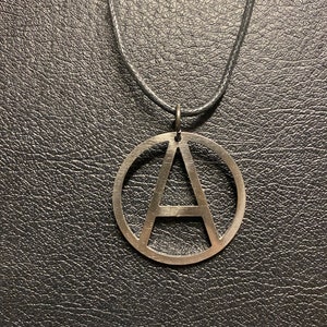 Op de afbeelding: Zilverkleurige hangerketting met de letter "A" in een cirkel. De hanger hangt aan een dun zwart koord. De achtergrond is een donker, getextureerd oppervlak, dat contrasteert met de zilveren hanger.