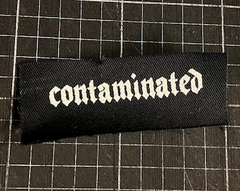 May include: A black fabric label with the word "contaminated" in white gothic lettering. The label is rectangular and appears to be made of a woven material. The background is a black grid.