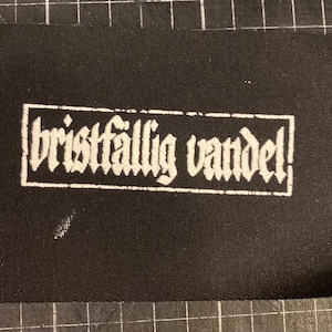 Op de afbeelding: Zwarte stoffen patch met witte tekst die "bristfällig vandel!" in een gotisch lettertype luidt.