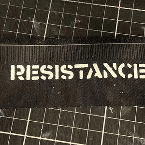 Könnte beinhalten: Schwarzer Stoff mit dem Wort "RESISTANCE" in weiß gedruckt.