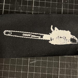 May include: Black fabric patch featuring a white chainsaw graphic with the word "CHAOS" printed on the blade. The patch is rectangular and has a stitched border, suitable for sewing onto clothing or accessories.