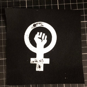 May include: Black square patch featuring a white feminist symbol, a fist inside a circle above a cross. The design has a distressed, vintage look. The patch is likely made of fabric and is suitable for sewing or ironing onto clothing or accessories.